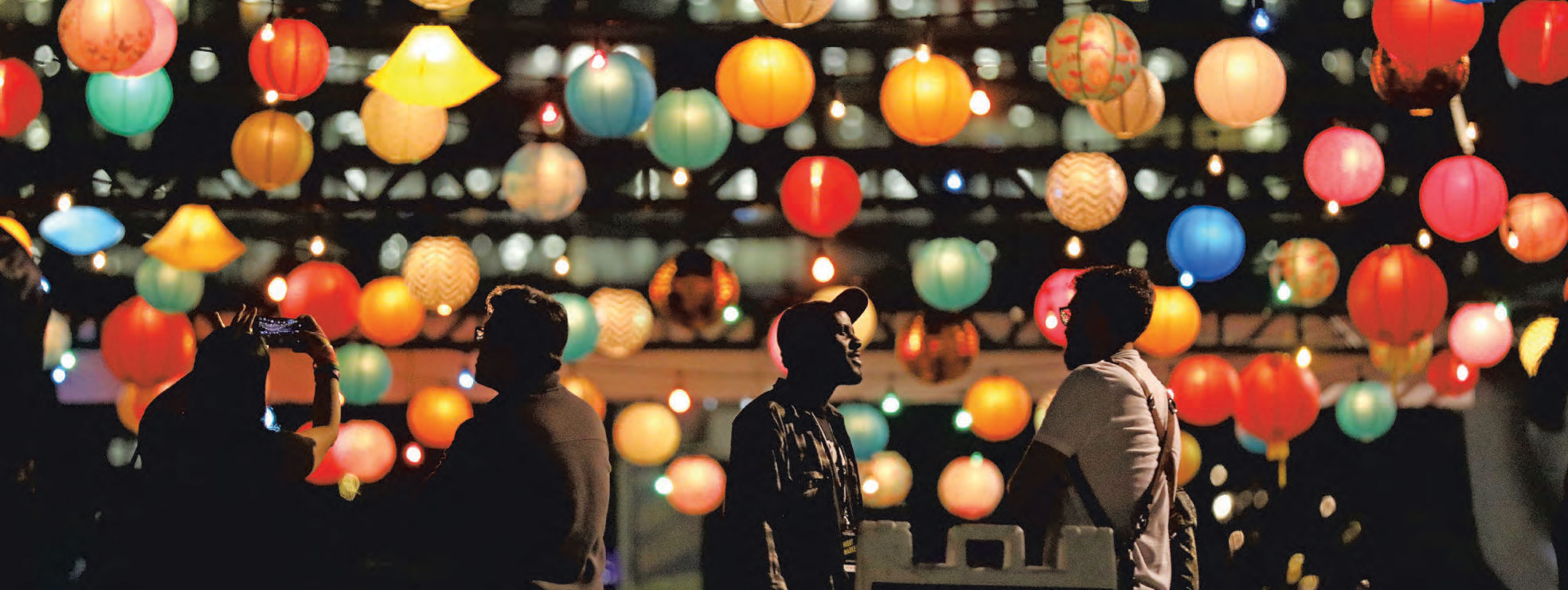 A casual group of people talking with each other in the evening, underneath colorful lanterns strung across an area outdoors.